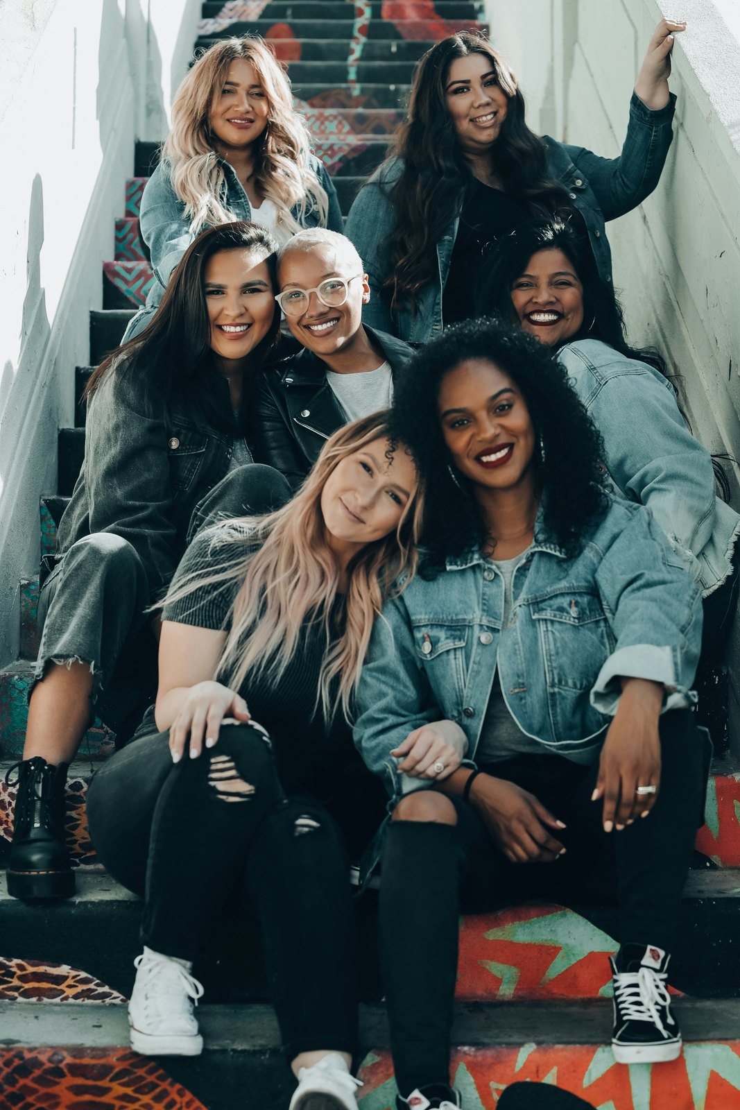 A group of seven women of diverse ages, skin tones, and hair styles sitting close together on steps, smiling and at ease with one another.